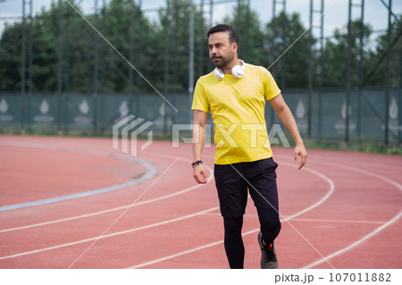 Bearded man in yellow t-shirt with wireless headphones standing on empty running track with red surface and white marking at contemporary sports ground in urban park Bearded man in yellow t-shirt with wireless headphones standing on empty running track with red surface and white marking at contemporary sports ground in urban park 107011882