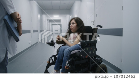 Hospital corridor: Woman with SMA in wheelchair talks to doctor 107012462