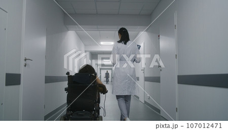 Female doctor walks down the clinic hallway, consults woman with disability 107012471