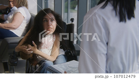 Woman with SMA in wheelchair talks to doctor in hospital lobby 107012479
