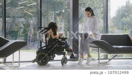 Female doctor consults patient with physical disability in clinic lobby Female doctor consults patient with physical disability in clinic lobby 107012499