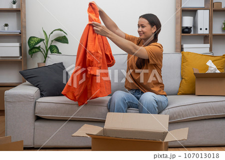 woman unpacks her clothes on her sofa, celebrating her express delivery and e-commerce fashion shopping experience.  107013018