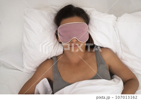 The tranquil lady, masked for sleep, peacefully rests on her pillow beneath a cozy blanket, a symbol of her healthy sleep regimen.  107013162