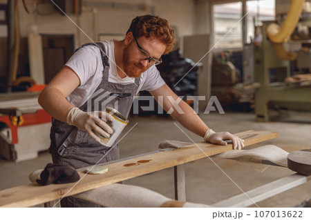 Craftsman coating a wooden board with protective oil in a workshop. Process of making a wood table Craftsman coating a wooden board with protective oil in a workshop. Process of making a wood table 107013622