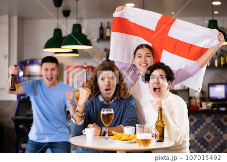 Enthusiastic company of young adult sports fans waving flag of England and supporting national team with beer in pub 107015492