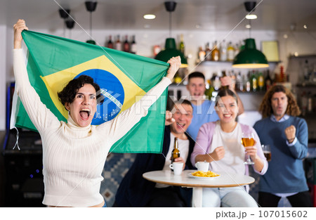 Company of emotional young adult fans supporting sports team of Brazil with state flag while resting with beer in bar 107015602