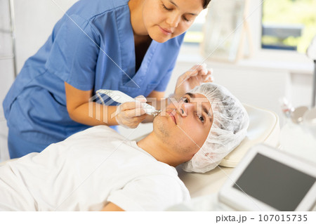 Young female cosmetologist doing microcurrent procedure to young man 107015735