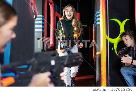 Joyous girl with laser pistol playing laser tag with friends on labyrinth Joyous girl with laser pistol playing laser tag with friends on labyrinth 107015850