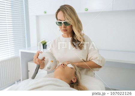 Portrait of certified cosmetologist wearing protective glasses conducting facial rejuvenation using modern apparatus for woman client. Professional beautician making face skin therapy for lifting. 107022126