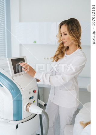 Vertical shot of professional female cosmetologist adjusts device for photorejuvenation using touchscreen in beauty clinic. Concept of non-surgery cosmetology, professional skincare, beauty treatment. Vertical shot of professional female cosmetologist adjusts device for photorejuvenation using touchscreen in beauty clinic. Concept of non-surgery cosmetology, professional skincare, beauty treatment. 107022161