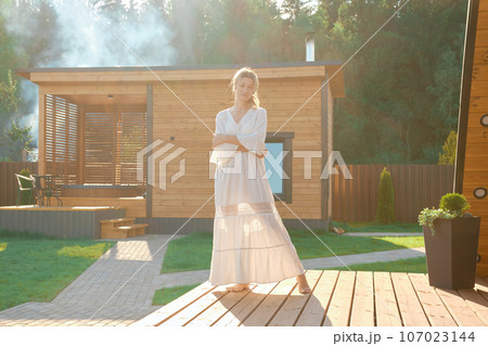 Dreamy woman in long white dress standing on terrace with hot tub and sauna on background 107023144