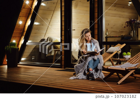 As evening falls, a woman wraps herself in a plaid and reads a book on the terrace As evening falls, a woman wraps herself in a plaid and reads a book on the terrace 107023182