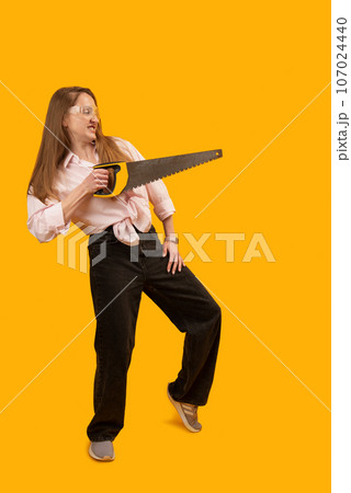 Young female model with hand saw posing for photo in studio against yellow background. Strong modern woman with man tools. Vertical frame. Copy space. 107024440