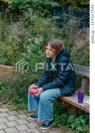 Blonde girl holding cup sitting on bench in the park. Autumn season. Young Teen Girl relaxing on bench with cup of coffee or tea. 107027544