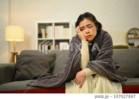 Sick woman suffering from strong headache or migraine while covered with a blanket sitting on sofa Sick woman suffering from strong headache or migraine while covered with a blanket sitting on sofa 107027867