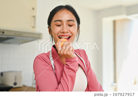 Charming young woman in sportswear eating an apple after exercising at home. Healthy food concept 107027917