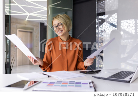 Joyful and successful businesswoman financier happy with achievement result, celebrating victory at workplace inside office, female worker holding contract reports and bills in hands. Joyful and successful businesswoman financier happy with achievement result, celebrating victory at workplace inside office, female worker holding contract reports and bills in hands. 107028571