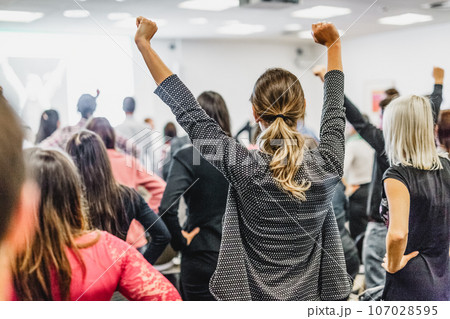 Life coaching symposium. Speaker giving interactive motivational speech at business workshop. Rear view of unrecognizable participants feeling empowered and motivated, hands raised high in air. 107028595