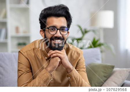 Close-up portrait of young smiling hispanic man smiling and looking at camera while sitting on sofa in living room. 107029568