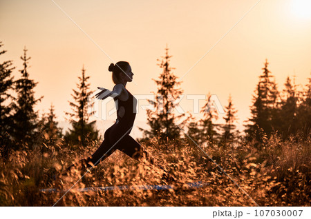 Attractive woman doing warrior one pose in the morning. Meditation in mountains at sunrise. Workout at fresh air outdoors. 107030007