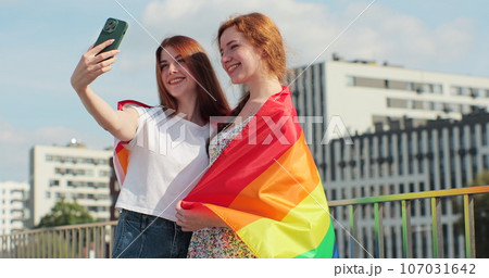 Lesbian couple taking selfie in the city. Two happy girlfriends pose with rainbow flag. Lgbt woman, showing love to each other. Happy lesbian relationships. 107031642