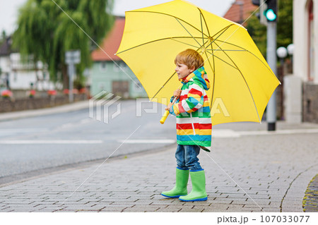 Little toddler boy playing with big yellow umbrella on rainy day. Happy positive child running through rain and puddles. Kid with rain clothes and rubber boots. Children activity on bad weather day. 107033077