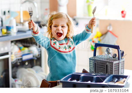 Cute little toddler girl helping in the kitchen with dish washing machine. Happy healthy blonde child sorting knives, forks, spoons, cutlery. Baby having fun with helping housework mother and father. 107033104