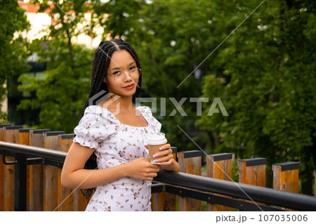 African young attractive woman in glasses stands on a bridge with a paper cup of coffee on the street during the day in the park in summer and looks at the camera African young attractive woman in glasses stands on a bridge with a paper cup of coffee on the street during the day in the park in summer and looks at the camera 107035006