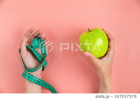 woman's one hand holds a green apple and the other hand holds a measuring tape, on a pink background. Weight loss and diet concept woman's one hand holds a green apple and the other hand holds a measuring tape, on a pink background. Weight loss and diet concept 107037379