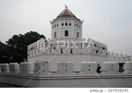 A white fort-like structure with a red roof, surrounded by a wall with black crosses, stands against a backdrop of trees and a clear sky. 107039245