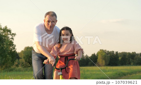 Happy father supports bicycle while little girl learning to ride in green field Happy father supports bicycle while little girl learning to ride in green field 107039621