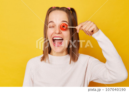 Funny joyful woman with ponytail in jumper holding lollipop isolated over yellow background covering eyes with sugary candy laughing expressing happiness. Funny joyful woman with ponytail in jumper holding lollipop isolated over yellow background covering eyes with sugary candy laughing expressing happiness. 107040190