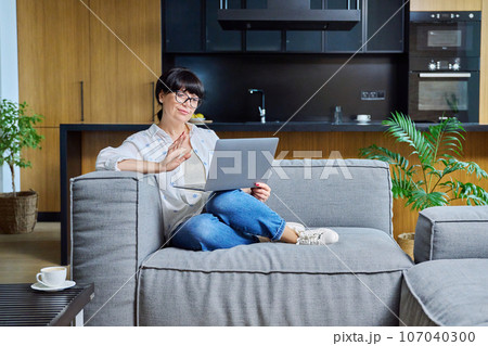 Mature positive woman sitting on sofa using laptop for video call 107040300