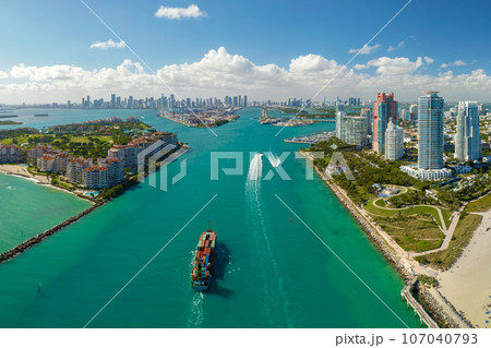 Aerial view of large container ship entering in Miami harbor main channel near South Beach. Luxurious hotels and apartment buildings on waterfront and high skyscrapers of downtown district in distance Aerial view of large container ship entering in Miami harbor main channel near South Beach. Luxurious hotels and apartment buildings on waterfront and high skyscrapers of downtown district in distance 107040793