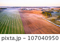 Warm sunny September morning. Autumn beautiful fog landscape. Small river curves in trees. Bright sunny misty riverside. Scenic view of rural sunrise. Village in Agriculture fields. 107040950
