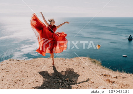 Woman in red dress on sea. Side view a Young beautiful sensual woman in a red long dress posing on a rock high above the sea on sunset. Girl on the nature on blue sky background. Fashion photo. 107042183