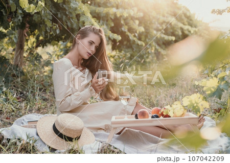 Woman picnic vineyard. Happy woman with a glass of wine at a picnic in the vineyard, wine tasting at sunset and open nature in the summer. Romantic dinner, fruit and wine. 107042209