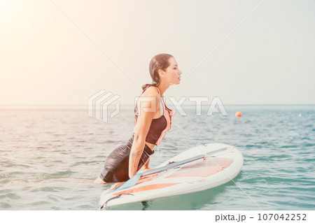 Sea woman sup. Silhouette of happy young woman in pink bikini, surfing on SUP board, confident paddling through water surface. Idyllic sunset. Active lifestyle at sea or river. 107042252