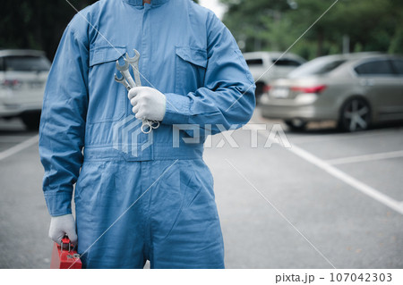 Close-up of a professional car mechanic's hands, wearing a blue work uniform and holding a large wrench while repairing a vehicle. Midsection shot. Close-up of a professional car mechanic's hands, wearing a blue work uniform and holding a large wrench while repairing a vehicle. Midsection shot. 107042303