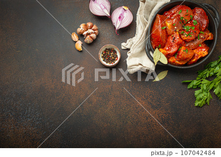 Cooking hearty meat stew. Raw uncooked chopped pieces of meat marinated with seasonings and parsley in black casserole dish top view, ingredients on dark rustic background. Space for text Cooking hearty meat stew. Raw uncooked chopped pieces of meat marinated with seasonings and parsley in black casserole dish top view, ingredients on dark rustic background. Space for text 107042484