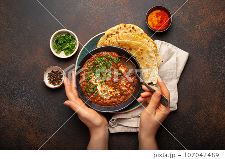 Female hands holding a bowl and eating traditional Indian Punjabi dish Dal makhani with lentils and beans served with naan flat bread, fresh cilantro on brown concrete rustic table top view 107042489