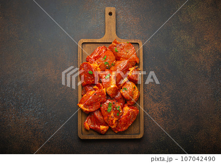 Raw uncooked chopped pieces of pork marinated with seasonings and parsley on wooden cutting board top view on dark rustic background. Cooking meal with marinated pork fillet pieces 107042490