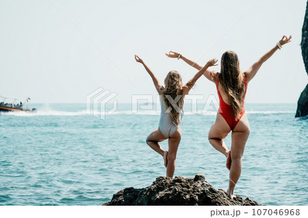 Woman and her daughter practicing balancing yoga pose on one leg up together on rock in the sea. Silhouette mother and daughter doing yoga at beach 107046968