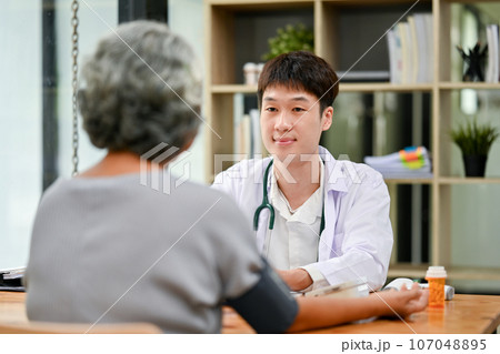 A professional Asian male doctor is diagnosing the disease and consulting with an old lady. 107048895