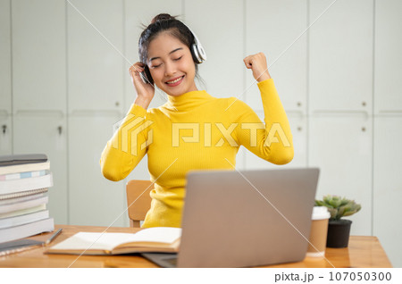 A cheerful Asian female college student enjoys listening to music on her headphones in a library. 107050300