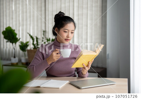 A beautiful Asian woman is reading a book while having her morning coffee calmly at a table. A beautiful Asian woman is reading a book while having her morning coffee calmly at a table. 107051726