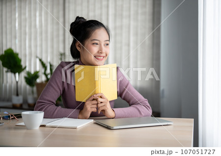 A beautiful and inspired Asian woman is daydreaming about happiness while enjoying reading a book. 107051727
