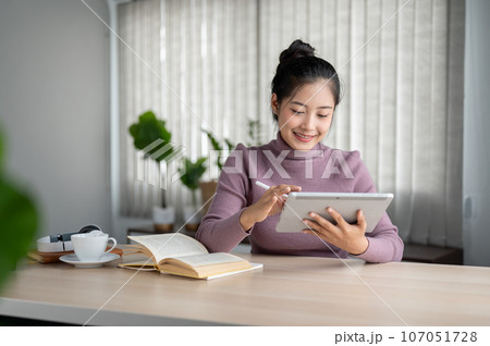 A happy and pretty Asian female college student using a digital tablet at a table in her living room 107051728