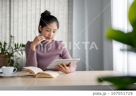 A focused Asian woman reads an online blog on her tablet, uses her digital tablet at a table. 107051729