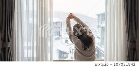 young woman in sweater looking through the window in winter season, happy female rising arms and stretching after waking at apartment or home in the morning. Lifestyle and Relaxing concepts young woman in sweater looking through the window in winter season, happy female rising arms and stretching after waking at apartment or home in the morning. Lifestyle and Relaxing concepts 107051942